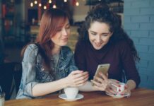 A woman with red hair is showing her phone to a woman with dark hair.