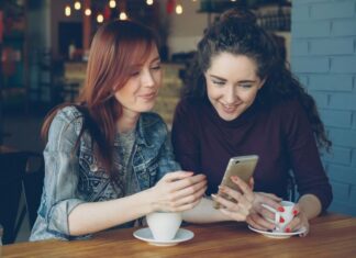 A woman with red hair is showing her phone to a woman with dark hair.