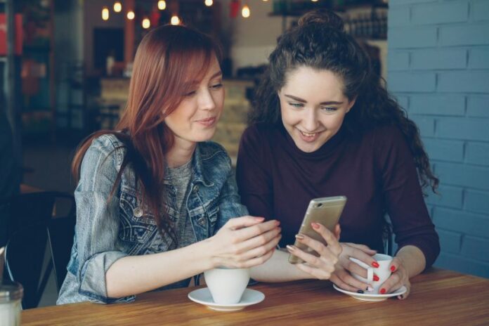 A woman with red hair is showing her phone to a woman with dark hair.