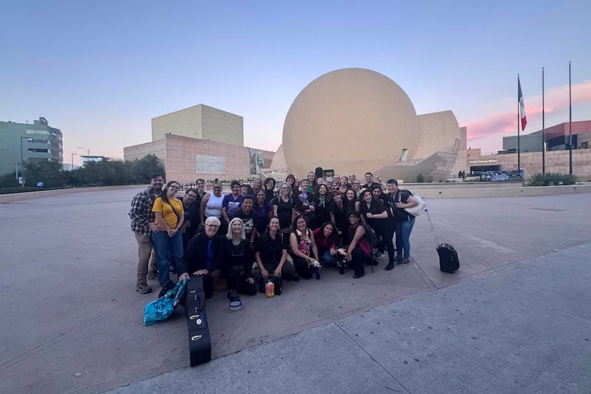 The Meraki Chorus of Tijuana, Baja California poses in an open plaza front of a round structure. In the background are other multistory buildings and a flagpole with a Mexican flag.