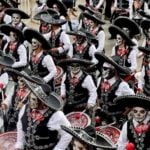 A large marching band performs in a Día de los Muertos parade in Mexico City, dressed in black mariachi-style outfits with sombreros and their faces painted as calaveras, or skulls.