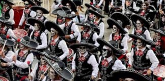 A large marching band performs in a Día de los Muertos parade in Mexico City, dressed in black mariachi-style outfits with sombreros and their faces painted as calaveras, or skulls.