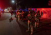 Roughly 20 Mexican soldiers stand on a street at night, bathed in the red and blue light of police cars