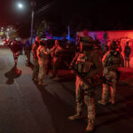 Roughly 20 Mexican soldiers stand on a street at night, bathed in the red and blue light of police cars