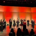 Women in black outfits typical of choir performances, each bearing a red flower on the lapel, perform onstage at the Tijuana Cultural Center. A female director leads the performers. The stage is illuminated by red and orange lighting.