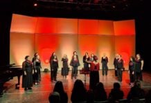 Voices Without Borders: How a Tijuana choir reached across the divide Women in black outfits typical of choir performances, each bearing a red flower on the lapel, perform onstage at the Tijuana Cultural Center. A female director leads the performers. The stage is illuminated by red and orange lighting.