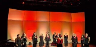 Women in black outfits typical of choir performances, each bearing a red flower on the lapel, perform onstage at the Tijuana Cultural Center. A female director leads the performers. The stage is illuminated by red and orange lighting.