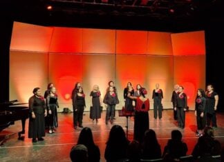 Voices Without Borders: How a Tijuana choir reached across the divide Women in black outfits typical of choir performances, each bearing a red flower on the lapel, perform onstage at the Tijuana Cultural Center. A female director leads the performers. The stage is illuminated by red and orange lighting.