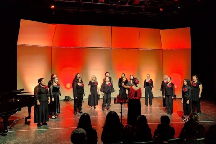 481470037_629979693117653_9126351746936436569_n Women in black outfits typical of choir performances, each bearing a red flower on the lapel, perform onstage at the Tijuana Cultural Center. A female director leads the performers. The stage is illuminated by red and orange lighting.