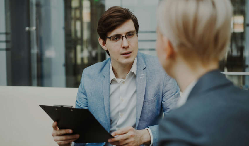A man wearing glasses and a light blue blazer holds a black clipboard while speaking to a woman with short blonde hair, whose back is partially to the camera. They appear to be in a professional setting, possibly an interview or meeting.