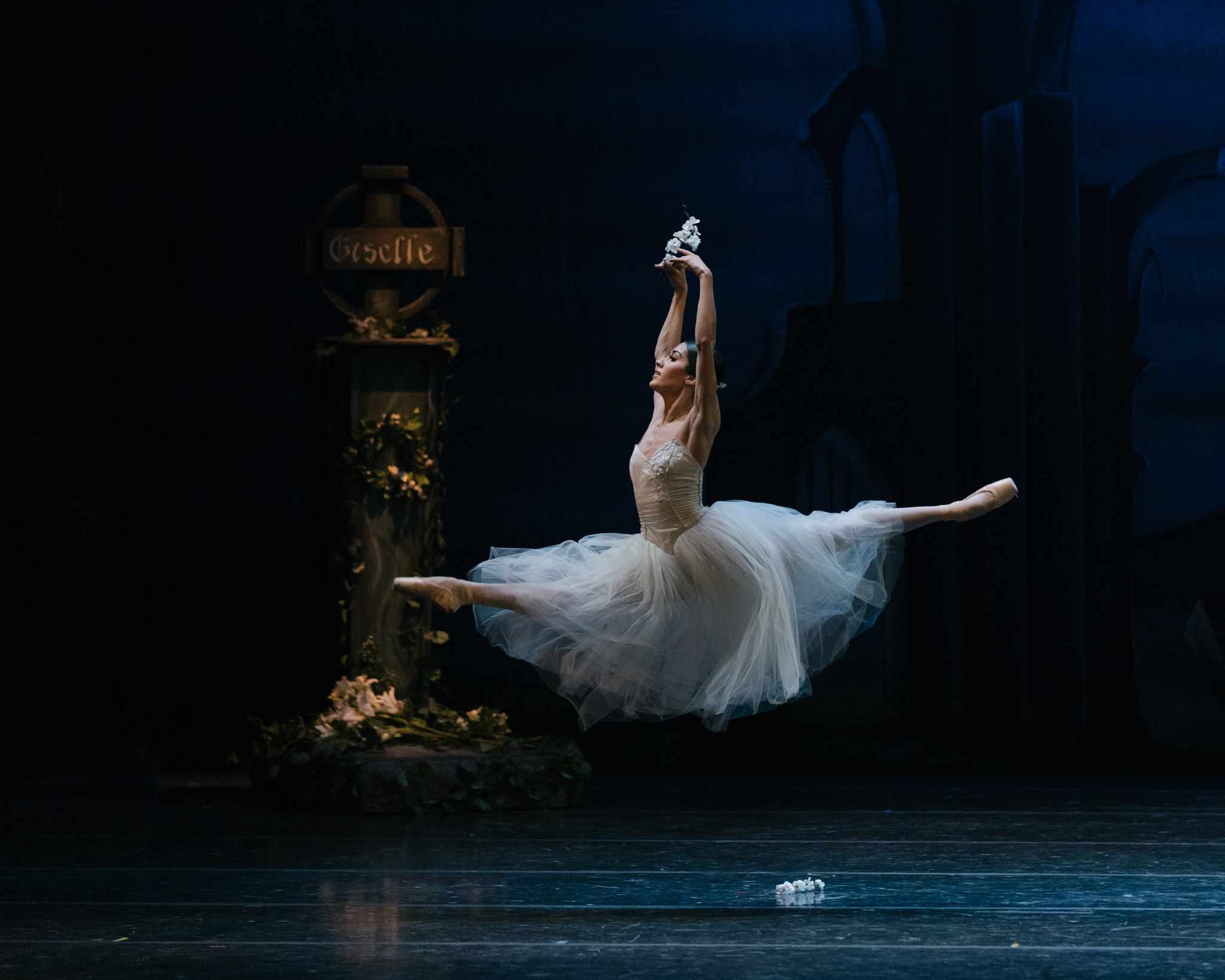 A ballerina successful  a achromatic  tutu performs a expansive  jeté crossed  a acheronian  stage, with a tombstone marked "Giselle" disposable   successful  the background.