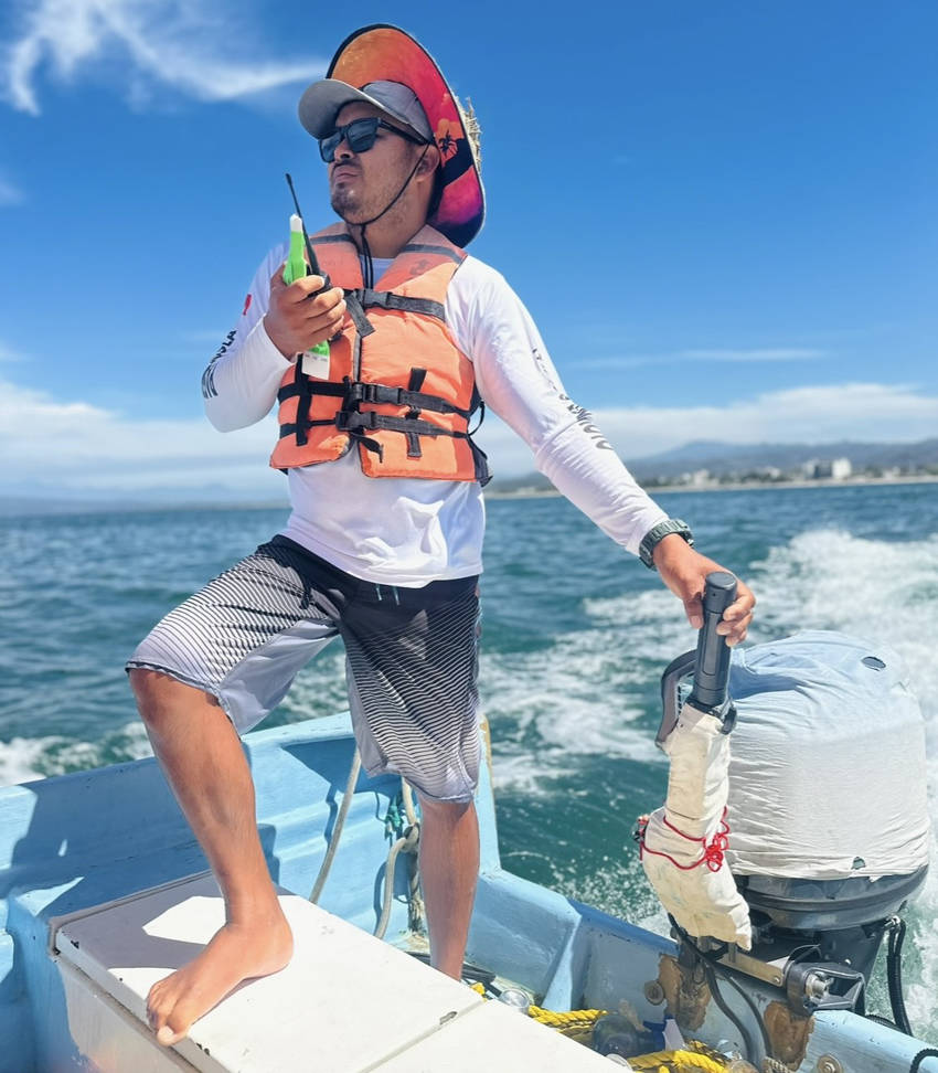 A man stands barefoot at the back of a small motorboat as it speeds across the water on a sunny day. He wears an orange life vest, a wide-brimmed sun hat over a baseball cap, and sunglasses. He holds a walkie-talkie and a green bottle in one hand while his other hand rests on the tiller of the outboard motor. The ocean and a distant coastline are visible in the background.