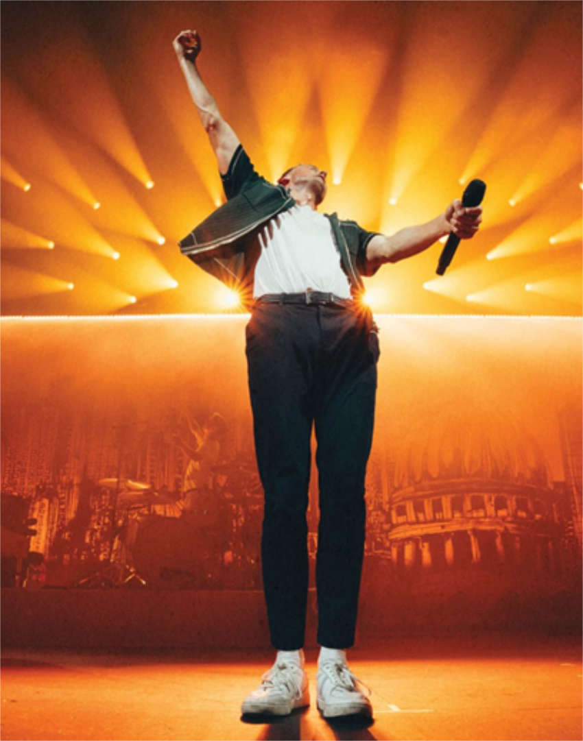 Full-length, low-angle photo of Tom Chaplin, lead singer of Keane, on stage during a concert. He stands facing the audience with his right arm raised in a fist of triumph and his left arm holding a microphone. He is wearing dark trousers, a white t-shirt, and a dark jacket, backlit by intense, golden-orange stage lights that fan out over the massive venue backdrop.