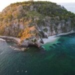 An aerial view of a small, rocky island covered in dense green vegetation. A secluded white-sand beach is nestled in a cove, and the island is surrounded by clear, emerald-green water that reveals dark coral reefs or rock formations beneath the surface.