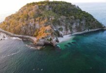 An aerial view of a small, rocky island covered in dense green vegetation. A secluded white-sand beach is nestled in a cove, and the island is surrounded by clear, emerald-green water that reveals dark coral reefs or rock formations beneath the surface.