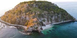 An aerial view of a small, rocky island covered in dense green vegetation. A secluded white-sand beach is nestled in a cove, and the island is surrounded by clear, emerald-green water that reveals dark coral reefs or rock formations beneath the surface.