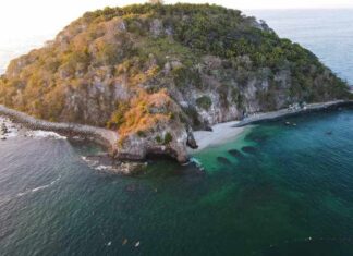 An aerial view of a small, rocky island covered in dense green vegetation. A secluded white-sand beach is nestled in a cove, and the island is surrounded by clear, emerald-green water that reveals dark coral reefs or rock formations beneath the surface.
