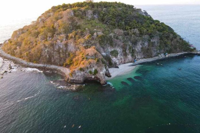 An aerial view of a small, rocky island covered in dense green vegetation. A secluded white-sand beach is nestled in a cove, and the island is surrounded by clear, emerald-green water that reveals dark coral reefs or rock formations beneath the surface.