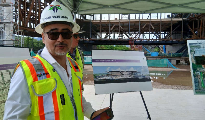 An older man with a goatee and sunglasses wearing a construction hard hat and a high-visibility safety vest stands at a construction site for the Puerto Vallarta International Airport's new Terminal 2. He is holding a smartphone and gesturing toward an easel with a rendering of the new airport terminal building, which is labeled "NUEVO EDIFICIO TERMINAL 2." The hard hat has a logo with the letters "PVR" and the words "PLAN 2025-2029" written on it.