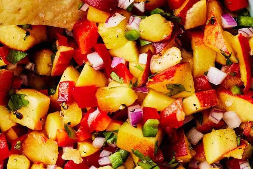 Close-up of vibrant, fresh peach salsa with diced yellow peaches, red bell peppers, red onion, and cilantro, served with a chip visible in the top left corner.