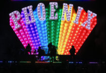 French rock band Phoenix performs on a dark stage in silhouette, backed by a massive, colorful LED screen displaying the band's name, PHOENIX, in large, marquee-style, light-up letters over a rainbow-colored, fan-shaped array of lights.