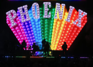 French rock band Phoenix performs on a dark stage in silhouette, backed by a massive, colorful LED screen displaying the band's name, PHOENIX, in large, marquee-style, light-up letters over a rainbow-colored, fan-shaped array of lights.