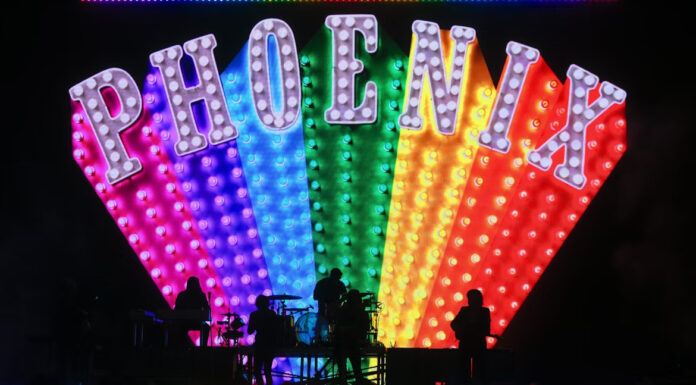 French rock band Phoenix performs on a dark stage in silhouette, backed by a massive, colorful LED screen displaying the band's name, PHOENIX, in large, marquee-style, light-up letters over a rainbow-colored, fan-shaped array of lights.