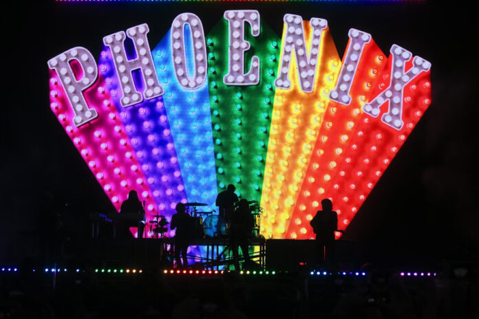 French rock band Phoenix performs on a dark stage in silhouette, backed by a massive, colorful LED screen displaying the band's name, PHOENIX, in large, marquee-style, light-up letters over a rainbow-colored, fan-shaped array of lights.
