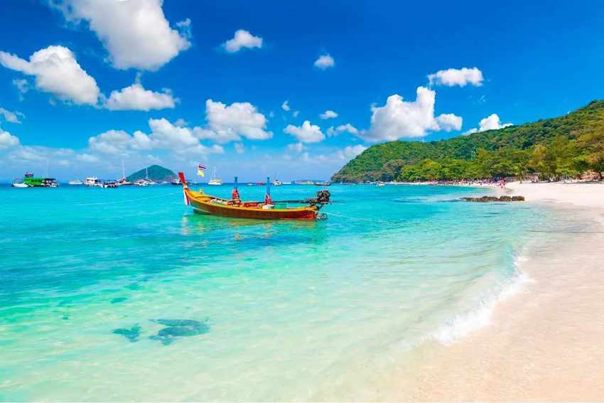 A traditional long-tail style wooden boat is anchored in calm, crystal-clear turquoise water just off a white-sand beach in Rincon de Guayabitos, Nayarit, Mexico. In the background, a lush, tree-covered hill slopes down to the sea, and other boats are visible in the distance. The scene is under a vibrant blue sky filled with puffy white clouds.