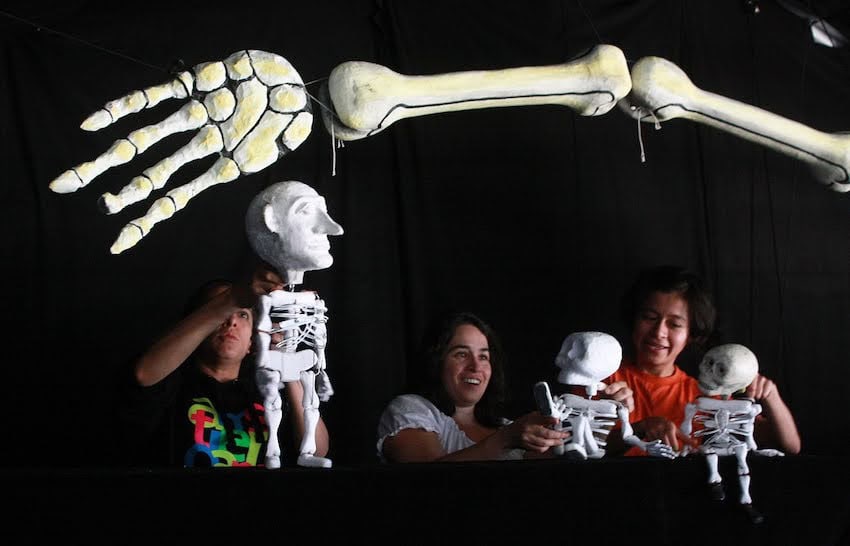 Three puppeteers run  respective  achromatic  papier-mâché skeleton puppets (calacas) of assorted  sizes down  a achromatic  stage, with a elephantine  skeletal limb  suspended supra  them.