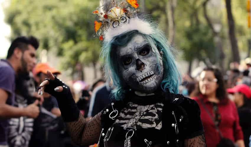 person with teal hairsbreadth  and elaborate skull look   overgarment   poses for the camera during a Día de los Muertos celebration, wearing a gothic-style outfit and a tiny  crown with skulls and marigolds.