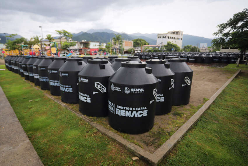 A low-angle photo shows dozens of new, black water storage tanks (likely Rotoplas or similar cisterns) arranged in rows on a grassy field in Puerto Vallarta, Jalisco, Mexico. The tanks are branded with logos for SEAPAL Vallarta and the text "CONTIGO SEAPAL RENACE" (With you, SEAPAL is reborn). In the background are buildings and green mountains under a cloudy sky.