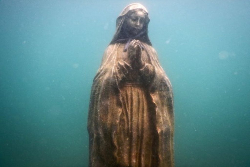 An underwater photograph shows a weathered statue of the Virgin Mary standing on the seabed. Her hands are clasped in prayer with a blue cord tied around them, and she is submerged in murky, turquoise-colored water.