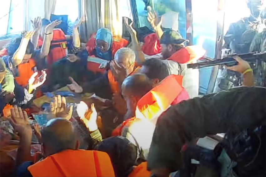 People in life vests on a small boat raise their hands in the air while a gun can be seen pointing at them from off camera