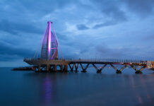 A long exposure night photo of the Muelle de Playa Los Muertos in Puerto Vallarta, Jalisco, Mexico. The iconic sail-like structure at the end of the pier is illuminated with a purple light against a blue twilight sky with dark clouds. The structure is connected to the shore by a long, dark pier extending over the calm ocean water.