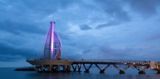 A long exposure night photo of the Muelle de Playa Los Muertos in Puerto Vallarta, Jalisco, Mexico. The iconic sail-like structure at the end of the pier is illuminated with a purple light against a blue twilight sky with dark clouds. The structure is connected to the shore by a long, dark pier extending over the calm ocean water.