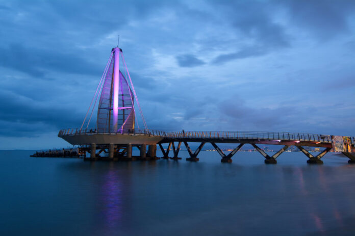 A long exposure night photo of the Muelle de Playa Los Muertos in Puerto Vallarta, Jalisco, Mexico. The iconic sail-like structure at the end of the pier is illuminated with a purple light against a blue twilight sky with dark clouds. The structure is connected to the shore by a long, dark pier extending over the calm ocean water.
