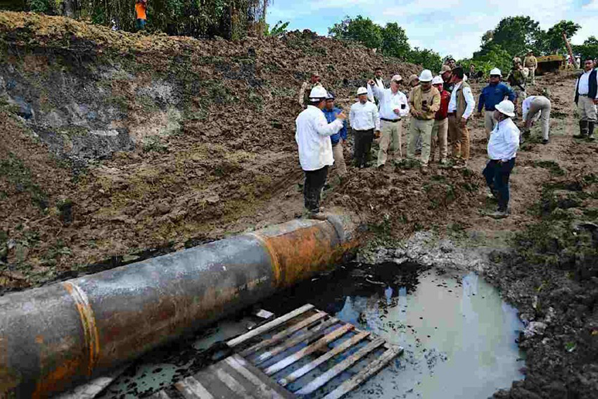 Engineers stand around an oil pipeline running along a muddy river bank