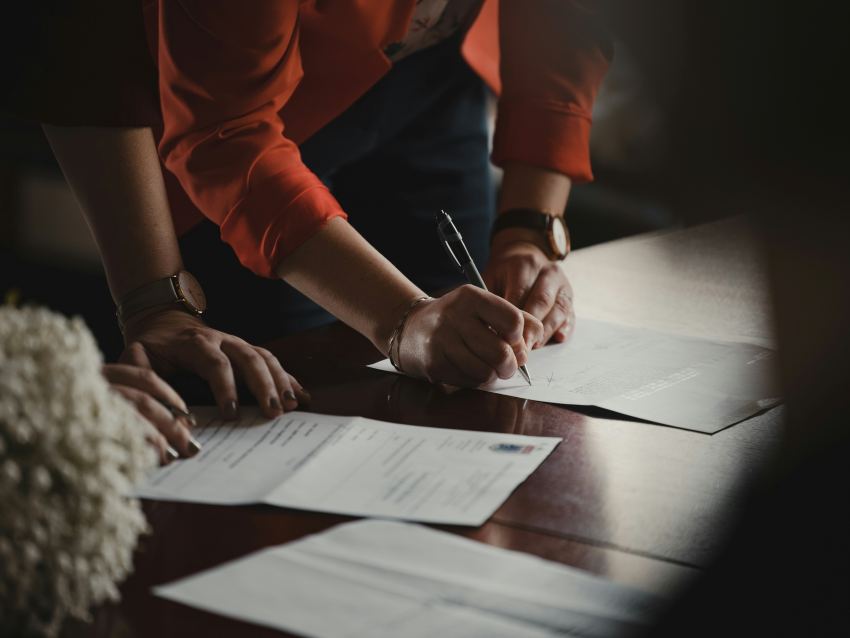 A view of a a woman's hands signing paperwork that's laid out on a wooden conference table. One paper has what looks like a governmental shield of an indeterminate country.