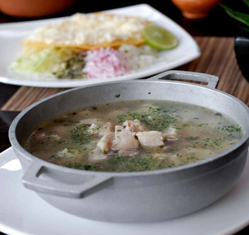 A metal bowl filled with traditional white pozole (clear broth, chunks of chicken or pork, and herbs) served alongside a side dish, perfect for a Dia de Muertos celebration.