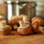 A close-up, shallow depth of field photo of several whole cremini mushrooms (baby portobello mushrooms), or champiñones, scattered across a wooden cutting board with a blurred stainless steel pot in the background.