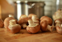 A close-up, shallow depth of field photo of several whole cremini mushrooms (baby portobello mushrooms), or champiñones, scattered across a wooden cutting board with a blurred stainless steel pot in the background.