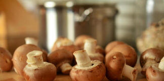 A close-up, shallow depth of field photo of several whole cremini mushrooms (baby portobello mushrooms), or champiñones, scattered across a wooden cutting board with a blurred stainless steel pot in the background.