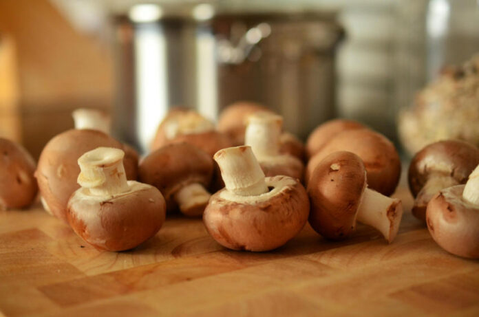 A close-up, shallow depth of field photo of several whole cremini mushrooms (baby portobello mushrooms), or champiñones, scattered across a wooden cutting board with a blurred stainless steel pot in the background.