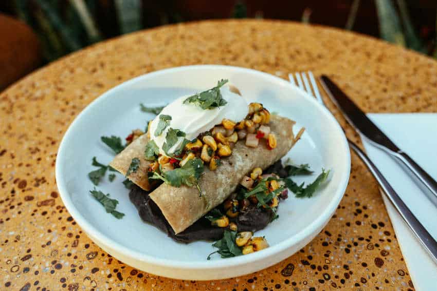 Spinach enchiladas on a bed of black beans, topped with roasted corn, red pepper, cilantro and crema, a delicious main course for a potential Dia de Muertos meal.