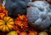 A staged close-up photo of a Día de Muertos altar featuring two types of Mexican pan de muerto. One is a large, light-colored loaf dusted heavily with white sugar, the other is dark but with the traditional pan de muerto "bones." Both are surrounded by vibrant orange cempasúchil marigold flowers and small orange and yellow decorative gourds.