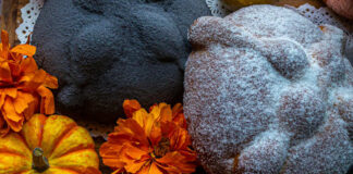 A staged close-up photo of a Día de Muertos altar featuring two types of Mexican pan de muerto. One is a large, light-colored loaf dusted heavily with white sugar, the other is dark but with the traditional pan de muerto "bones." Both are surrounded by vibrant orange cempasúchil marigold flowers and small orange and yellow decorative gourds.