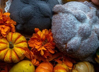 A staged close-up photo of a Día de Muertos altar featuring two types of Mexican pan de muerto. One is a large, light-colored loaf dusted heavily with white sugar, the other is dark but with the traditional pan de muerto "bones." Both are surrounded by vibrant orange cempasúchil marigold flowers and small orange and yellow decorative gourds.