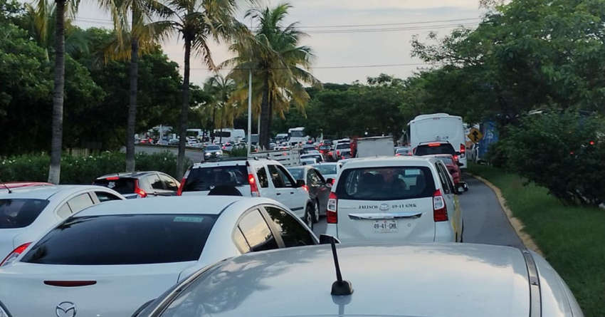 A photo taken from inside a car in heavy, slow-moving traffic on a road in Puerto Vallarta, Jalisco, Mexico, showing a long line of cars, including an ambulance and a bus, bumper-to-bumper. The road is lined with lush green tropical foliage and palm trees under an overcast sky.