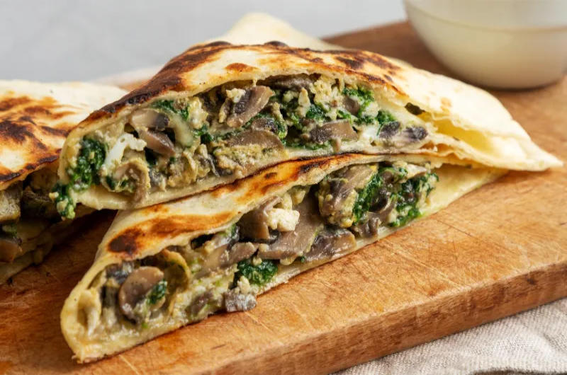 A close-up shot of a halved, folded, and pan-fried quesadilla resting on a wooden cutting board. The visible filling inside the flour tortilla is a savory mix of sliced brown mushrooms (champiñones), green leafy epazote and melted white cheese.